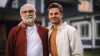 un grand-père et son fils ou petit fils souriant devant une maison floutée