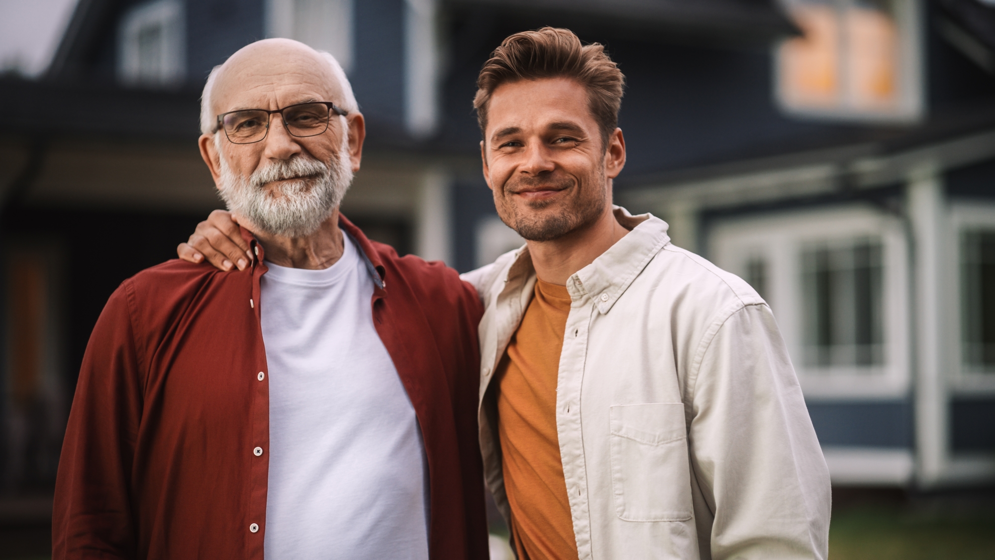 un grand-père et son fils ou petit fils souriant devant une maison floutée